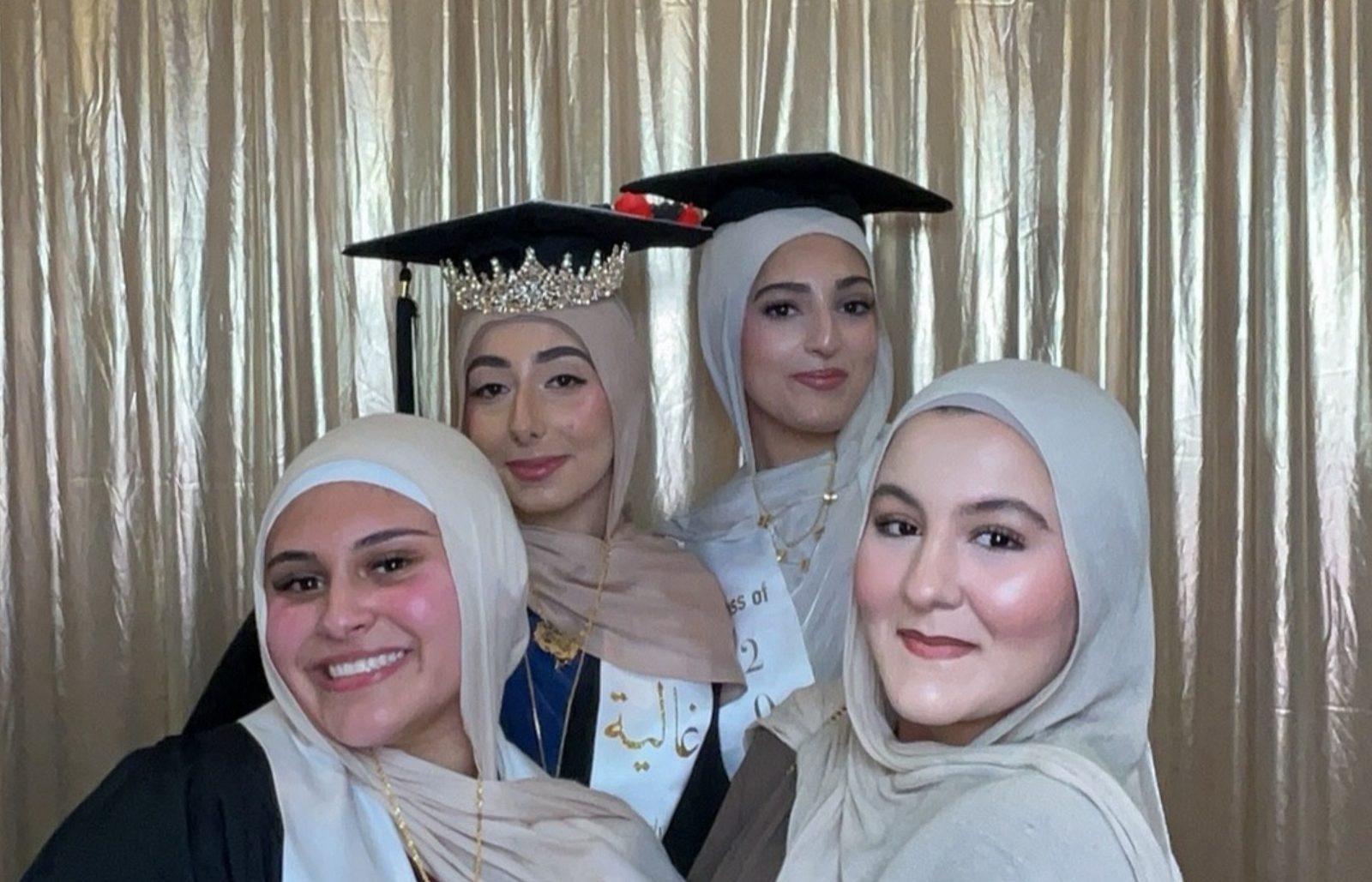 Four graduates in caps taking a group selfie in front of a gold backdrop