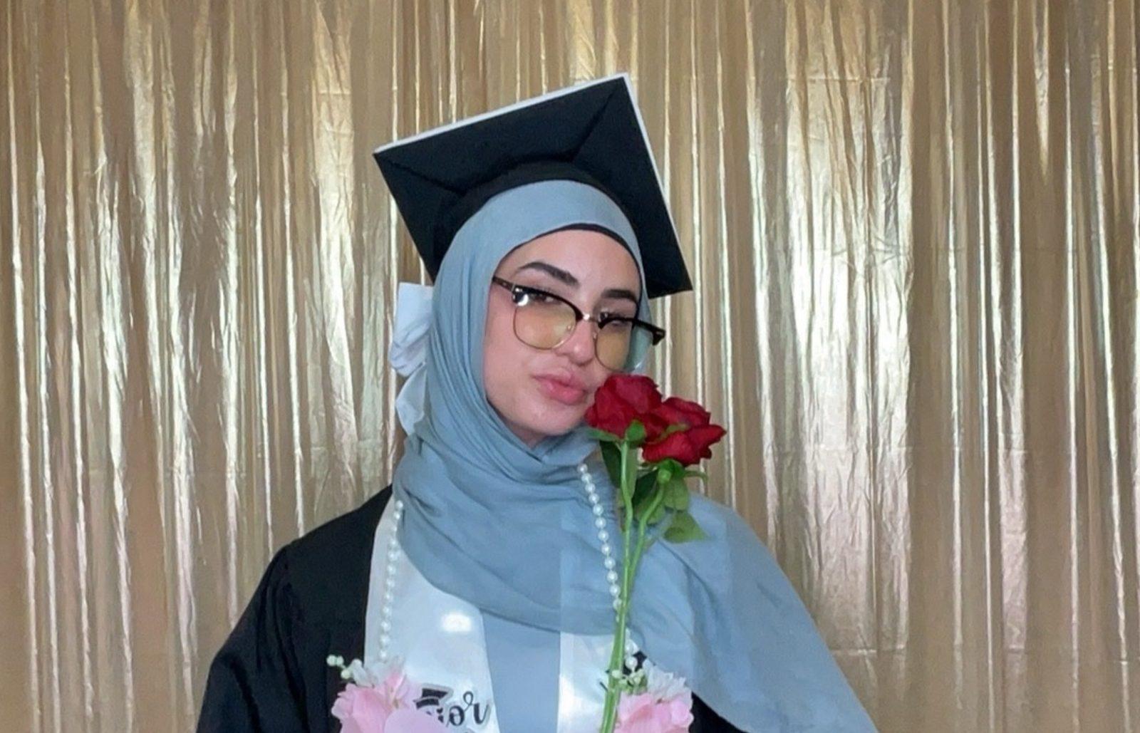 Graduate in cap and gown smelling red roses in front of a gold photo booth backdrop